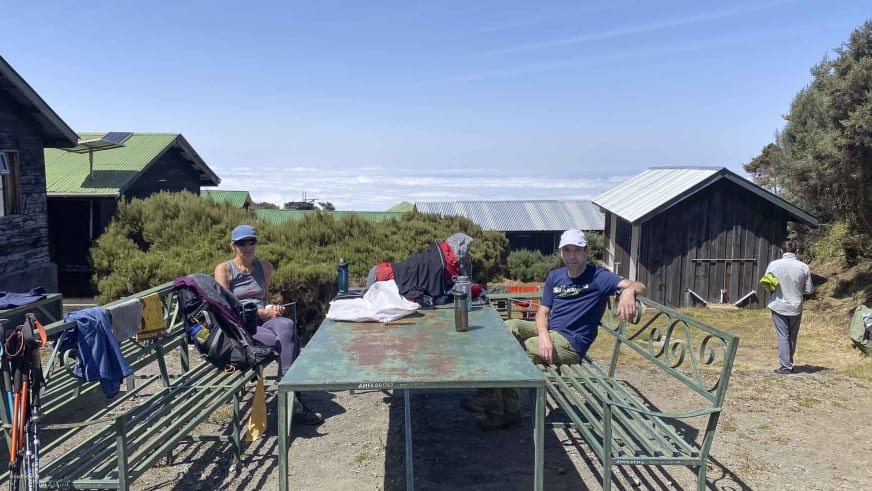 Vier Personen pausieren am Metalltisch der Sattelhütte (Mt. Meru) vor Stein- und Holzhütten.