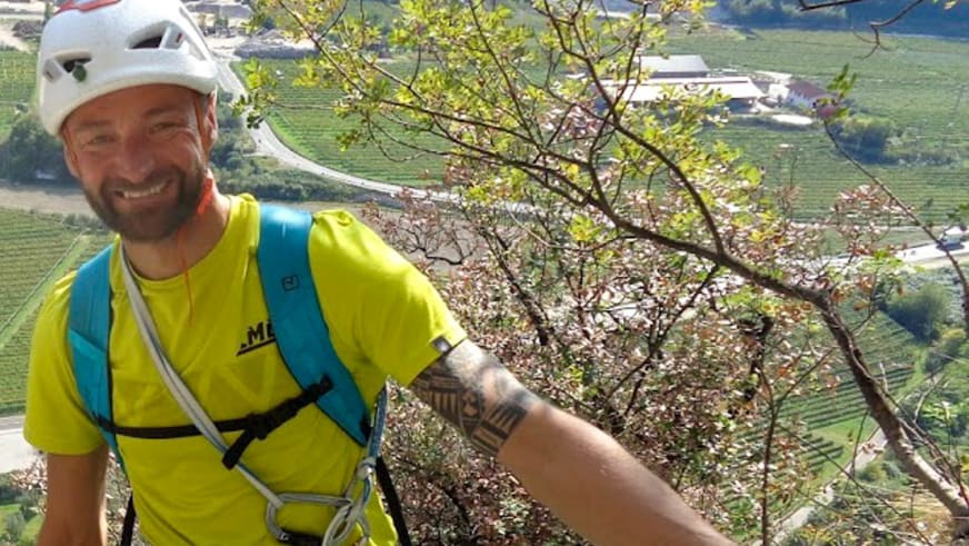 Bergführer Klaus Petz mit weißem Kletterhelm und gelbem T-Shirt lächelt vor terrassenartigen Weinbergen im Hintergrund.