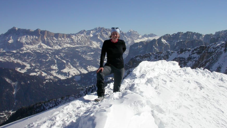 Auf einem verschneiten Gipfel sitzt Bergwanderführerin Birgit Eder mit weißer Mütze vor schneebedeckten Bergen.