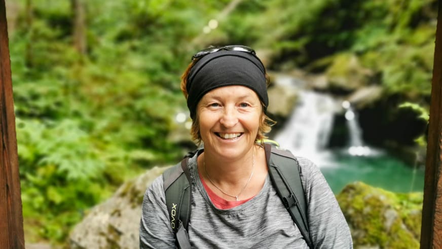 Bergwanderführerin Birgit Eder lächelt vor türkisgrünem Wasserfall mit schwarzer Mütze und grauem Wandershirt.