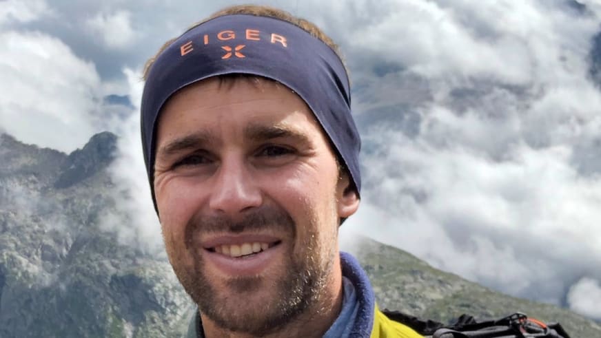 Benjamin Mäder trägt blaue Eiger-Mütze und blauen Fleecepullover, Bergwanderführer vor bewaldeten Bergen.