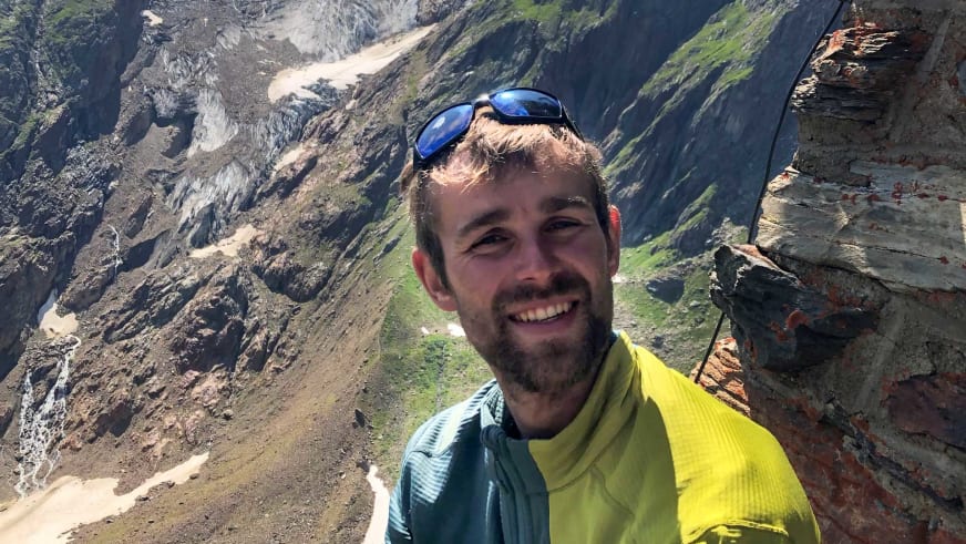 Bergwanderführer Benjamin Mäder trägt blaue Sonnenbrille, gelbes Shirt und türkisen Rucksack vor Berglandschaft.