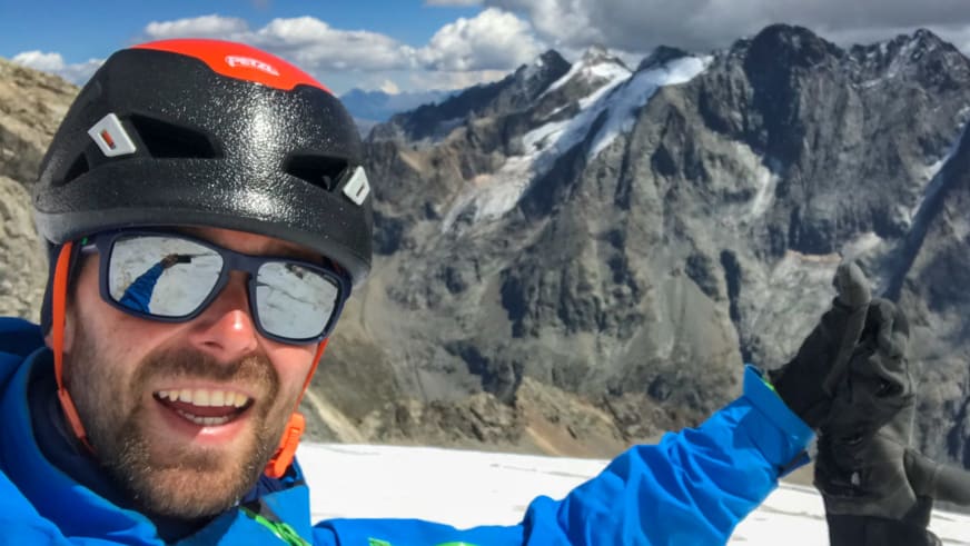 Bergwanderführer Benjamin Mäder macht ein Selfie im Schnee mit schwarzem Helm und blauer Jacke vor Bergpanorama.