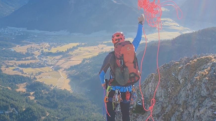 Bergwanderführer Dennis Brida hält rote Seile über dem Kopf und steht mit Klettergurt auf einem Felsvorsprung mit Blick ins Tal.