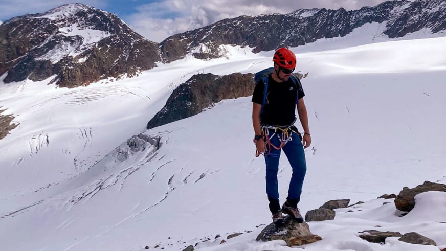 Bergwanderführer Dennis Brida steht mit rotem Helm und Klettergurt auf schneebedecktem Terrain vor einer Gletscherlandschaft.