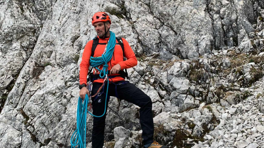 Bergwanderführer Dennis Brida sitzt mit rotem Helm und türkisem Kletterseil auf hellem Kalksteinfels in alpinem Gelände.