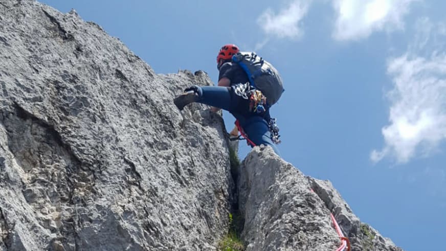 Bergwanderführer Dennis Brida klettert mit rotem Helm an einer steilen grauen Kalksteinwand gegen blauen Himmel.