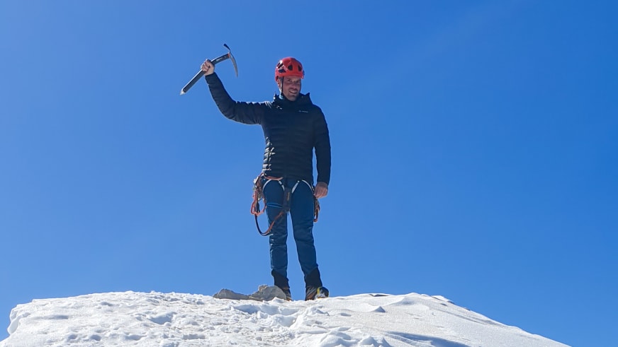 Bergwanderführer Dennis Brida hält triumphierend einen Eispickel hoch und trägt roten Helm auf einem verschneiten Gipfel vor blauem Himmel.