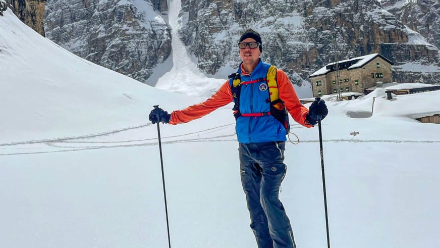 Bergführer Silvestro Franchini steht mit Skistöcken auf verschneiter Ebene vor steinerner Berghütte in blauer Skijacke.