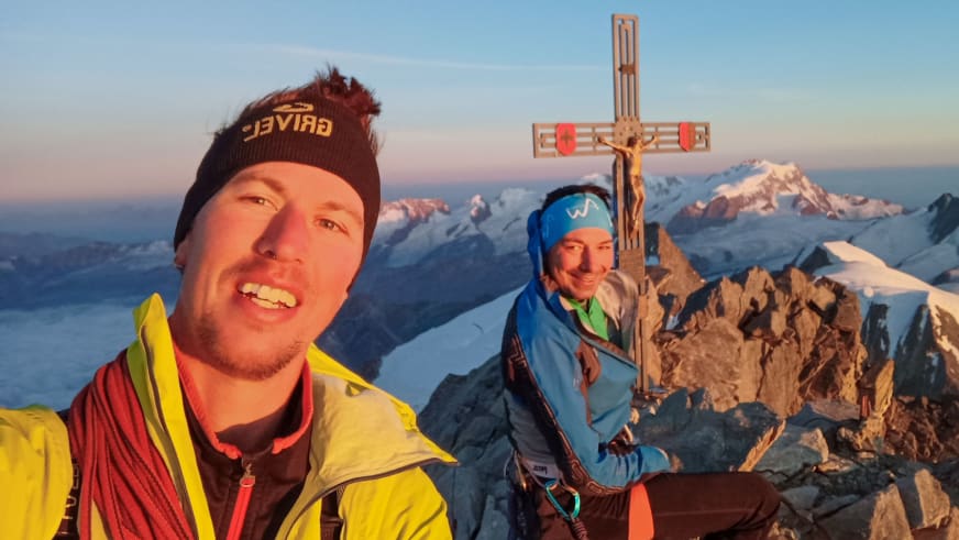 Zwei Bergsteiger auf Felsgipfel bei Sonnenaufgang, links Bergführer Silvestro Franchini in gelber Jacke neben hölzernem Gipfelkreuz.