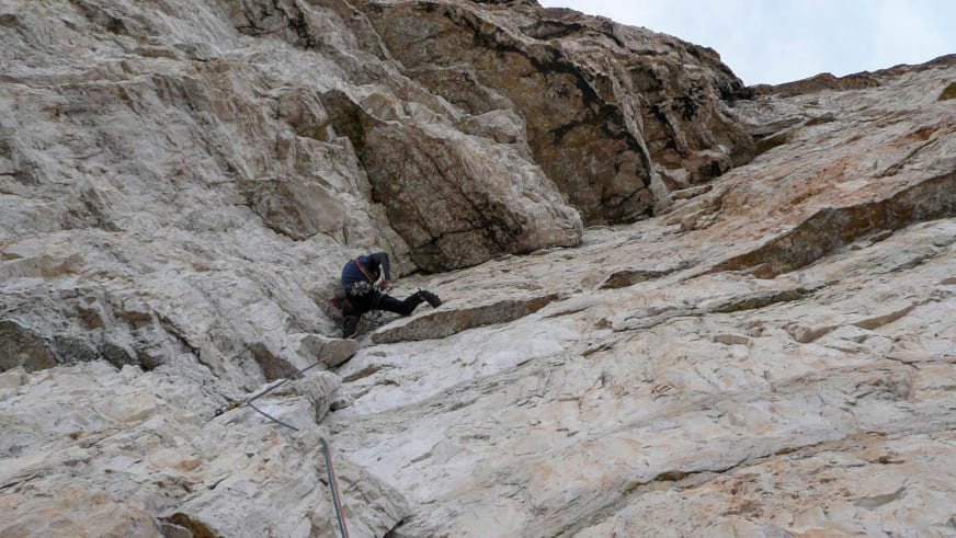 An einer hellen Kalksteinwand klettert Bergführer Klaus Karlstätter mit Helm und orangefarbenem Kletterseil zwischen Felsspalten.