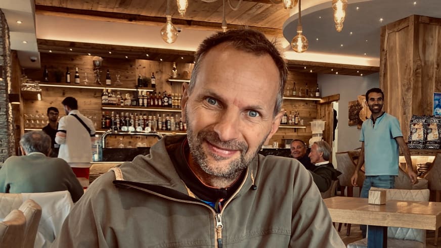 Bergführer Klaus Karlstätter lächelt in khakifarbener Fleecejacke im Vordergrund einer gemütlichen Berghütte mit Holzbalken und warmer Beleuchtung.