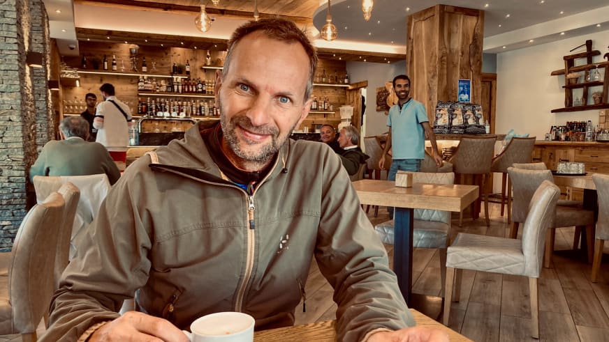Bergführer Klaus Karlstätter sitzt lächelnd in grauer Softshelljacke an einem Holztisch mit weißer Kaffeetasse vor sich in alpiner Hütte.