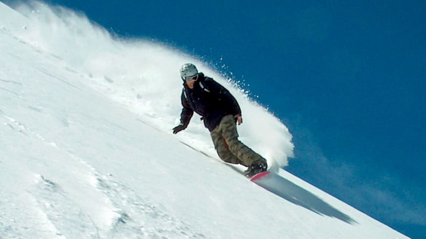 Bergführer Markus Arzberger fährt auf einem Snowboard einen steilen Schneehang hinab und wirbelt dabei Pulverschnee auf.