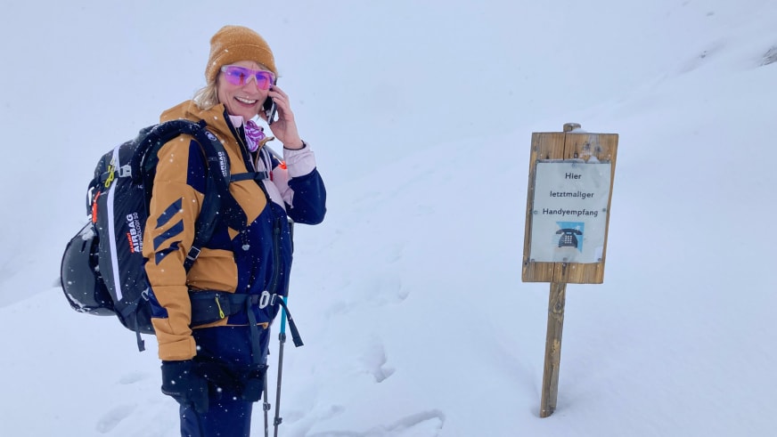 In blau-oranger Winterkleidung telefoniert Bergwanderführerin Stefanie Erdmann neben Holzschild im Schnee.