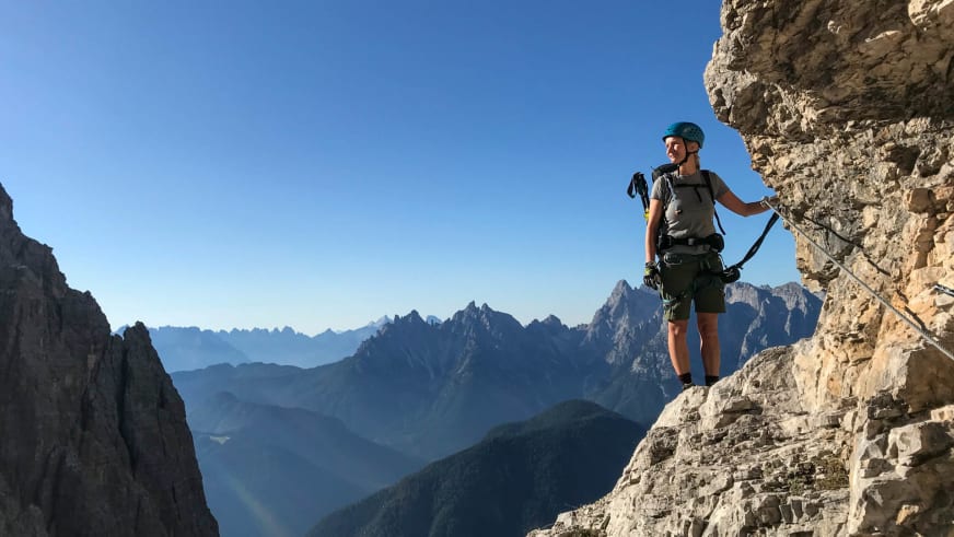 Bergwanderführerin Stefanie Erdmann steht mit türkisem Helm und Klettergurt auf Felsband vor Bergpanorama.