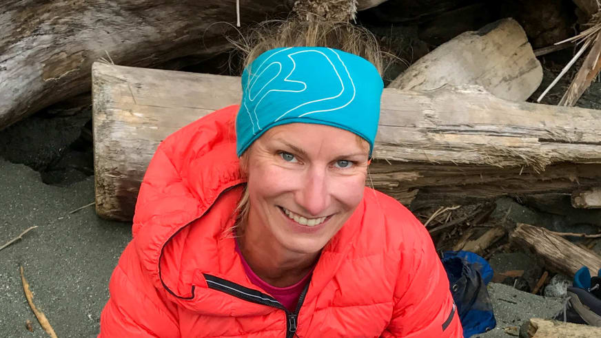 Portrait von Bergwanderführerin Stefanie Erdmann mit türkiser Mütze und roter Daunenjacke vor Holzstämmen.