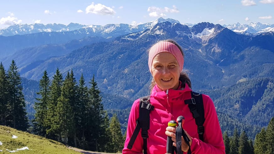Bergwanderführerin Julia Riedl lächelt mit Wanderstöcken und pinker Jacke auf grüner Almwiese vor schneebedeckten Gipfeln.