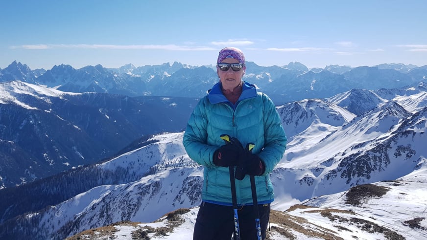 Bergwanderführerin Julia Riedl steht lächelnd in türkisfarbener Winterjacke auf schneebedecktem Gipfel vor Alpenpanorama.