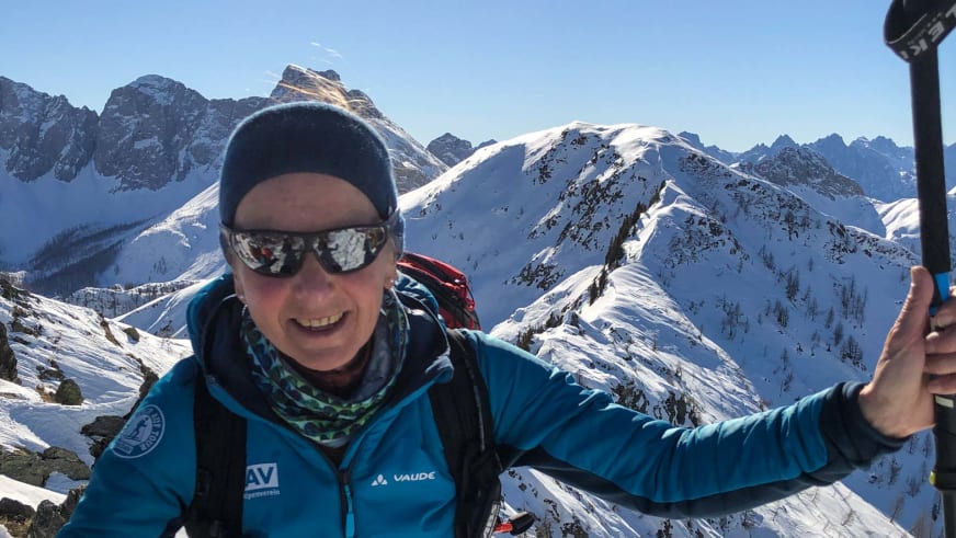 Bergwanderführerin Julia Riedl macht Selfie mit Selfie-Stick in blauer Winterjacke vor schneebedeckten Bergen.