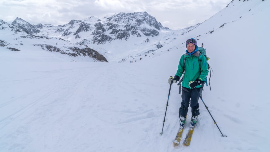 Bergwanderführerin Julia Riedl steht auf gelb-grünen Skiern in grüner Winterjacke vor verschneiter Hochgebirgslandschaft.