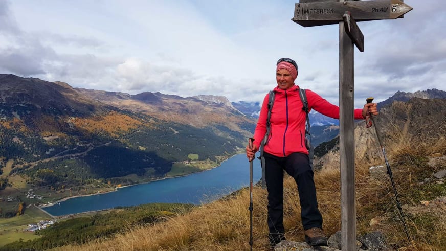 Bergwanderführerin Julia Riedl steht lächelnd neben Wegweiser zum Mittereck mit Wanderstöcken und roter Softshelljacke.