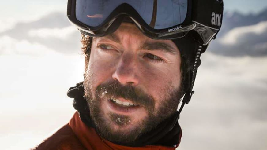 Bergführer Lukas Höllrigl lächelt in die Kamera mit Skibrille auf der Stirn und roter Jacke vor verschneiter Bergkulisse.