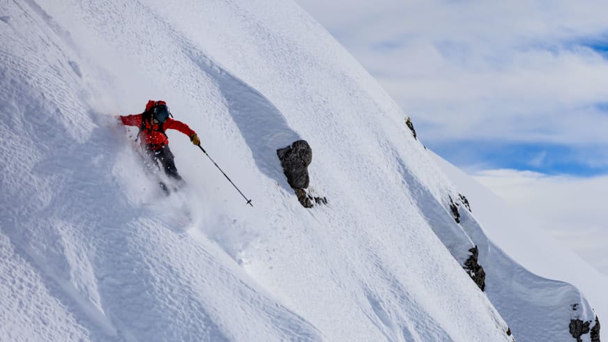 Auf steiler Abfahrt wirbelt Bergführer Lukas Höllrigl in roter Jacke eine große Schneewolke im Tiefschnee auf.