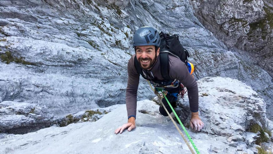 Bergführer Lukas Höllrigl mit grauem Helm lächelt in die Kamera während er an heller Kalksteinwand klettert.