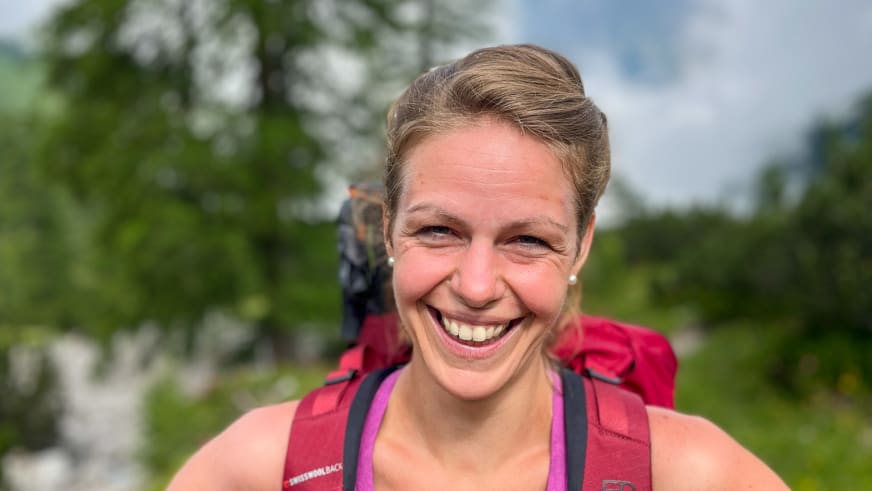 Portrait zeigt Bergwanderführerin Patricia Neumair lächelnd mit blonden Haaren und rotem Oberteil vor grünen Bäumen.