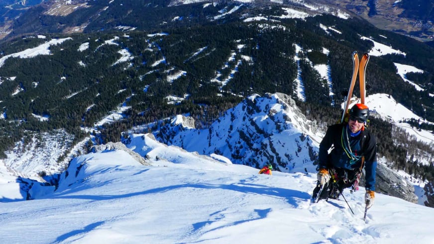 Auf einem verschneiten Gipfel steht Bergführer Lukas Seedoch mit orange Skiern auf dem Rücken befestigt.