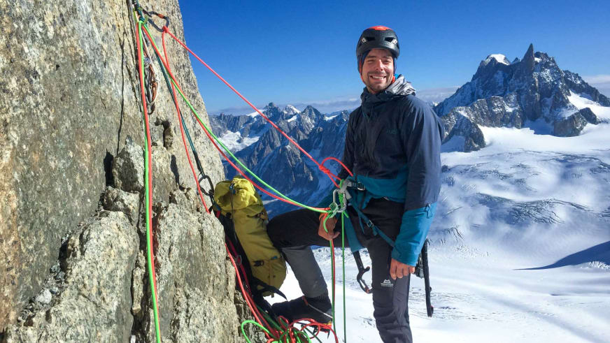 Bergführer Lukas Seedoch sitzt lächelnd im Klettergurt an einer Granitwand mit verschiedenfarbigen Kletterseilen.