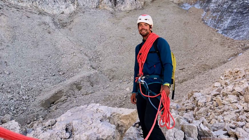Lächelnder Bergführer Lukas Seedoch mit weißem Helm und rotem Kletterseil in alpiner Gerölllandschaft.