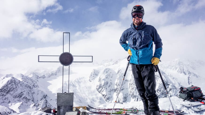 Bergführer Lukas Seedoch steht mit Skistöcken neben einem Metallkreuz auf einem verschneiten Gipfel.