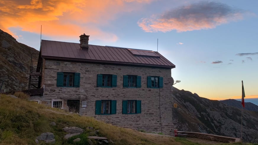 Das Rifugio Alfredo Rivetti mit türkisgrünen Fensterläden und italienischer Flagge während der goldenen Abenddämmerung.