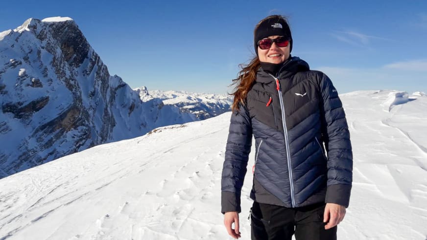 Mit grauer Daunenjacke und roter Sonnenbrille lächelt Bergwanderführerin Madlen Bucher auf schneebedecktem Berghang.