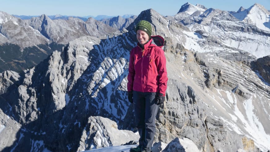 Bergwanderführerin Anna Bodenstedt steht lächelnd auf einem Gipfel mit roter Jacke und Rucksack vor Kalksteinbergen.