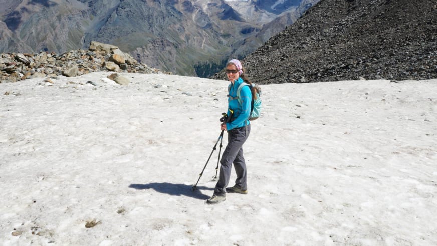 Bergwanderführerin Anna Bodenstedt steht mit Wanderstöcken auf einer Schneefläche in türkiser Jacke und rosa Mütze vor Hochgebirgslandschaft.