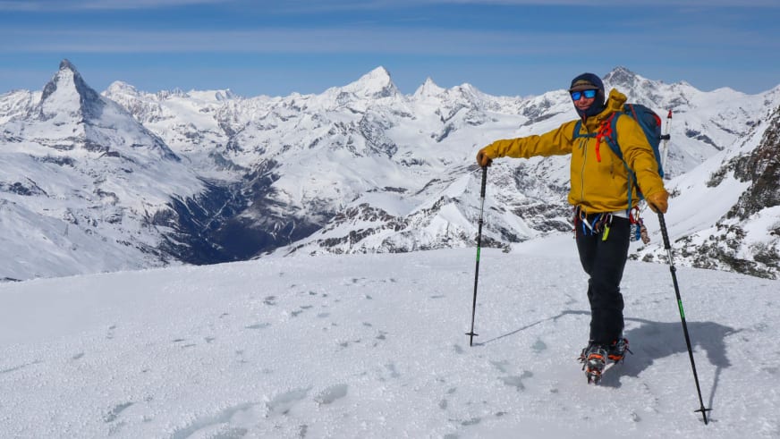 In gelber Winterjacke und mit Steigeisen steht Bergführer Jakob Fink auf verschneitem Hang vor Bergpanorama.