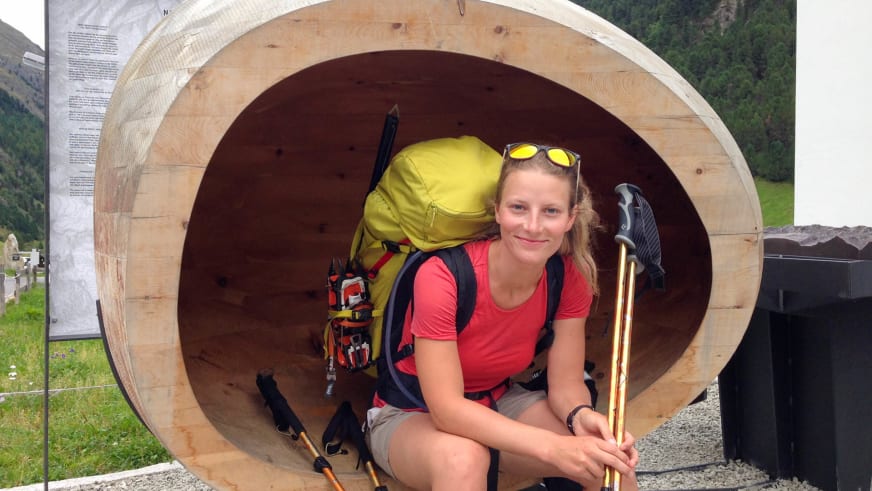 In einer hellen Holzröhre sitzt Bergwanderführerin Anna Biller mit gelbem Trekkingrucksack und blickt in die Kamera.
