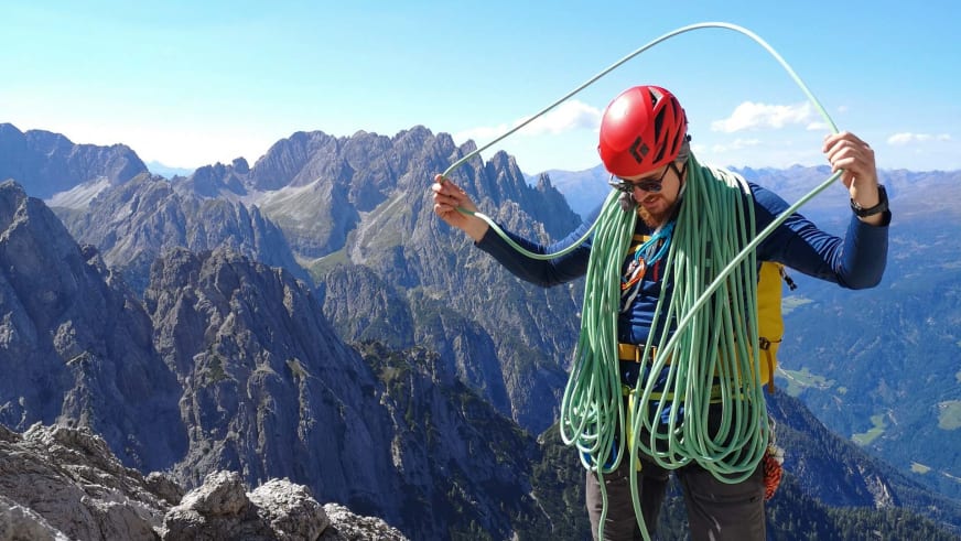Bergwanderführer Timo Reichert mit rotem Helm sortiert grünes Seil auf felsigem Gipfelbereich vor Bergpanorama.