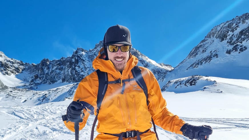 Bergwanderführer Timo Reichert lächelt mit Skistöcken in oranger Jacke vor verschneiter Gletscherlandschaft.
