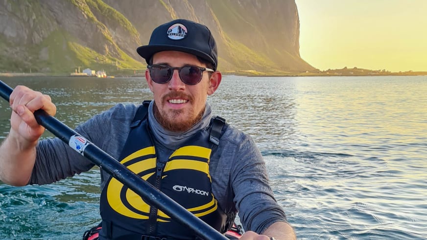Bergwanderführer Timo Reichert paddelt im Kajak mit gelb-schwarzer Schwimmweste vor grünen Berghängen.