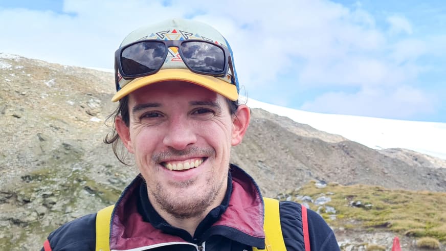 Nahaufnahme von Bergwanderführer Timo Reichert mit bunter Kappe vor weiter Berglandschaft mit schneebedeckten Gipfeln.