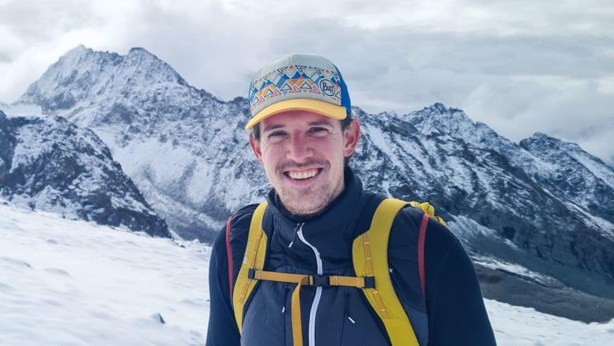 Bergwanderführer Timo Reichert lächelt in bunter Kappe und blauer Winterjacke vor schneebedeckten Bergen.