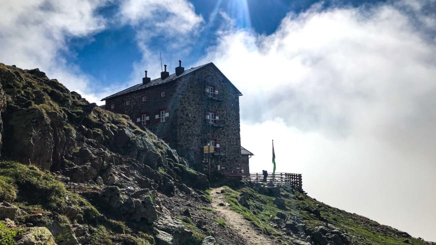 Die Breslauer Hütte aus Naturstein auf felsigem Bergkamm mit grün-weißen Fensterläden und Fahne in den Ötztaler Alpen.
