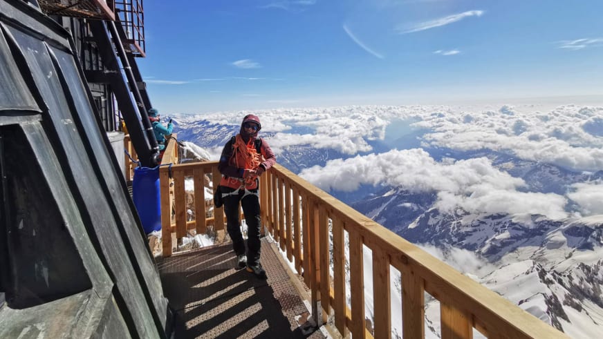 Auf einer Holzterrasse vor einem Berggebäude steht Bergführerin Elisabeth Seibt am Geländer vor schneebedeckten Bergen.