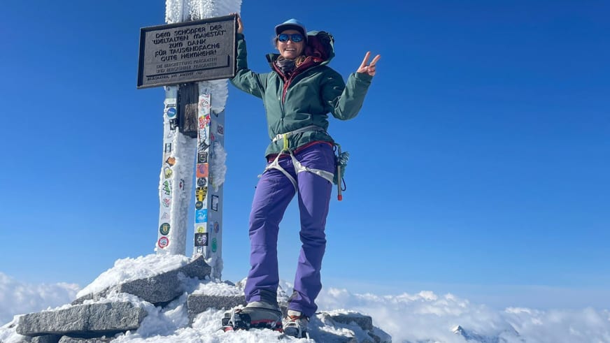 Bergführerin Elisabeth Seibt macht ein Victory-Zeichen neben einem mit bunten Aufklebern beklebten Gipfelkreuz auf schneebedecktem Gipfel.