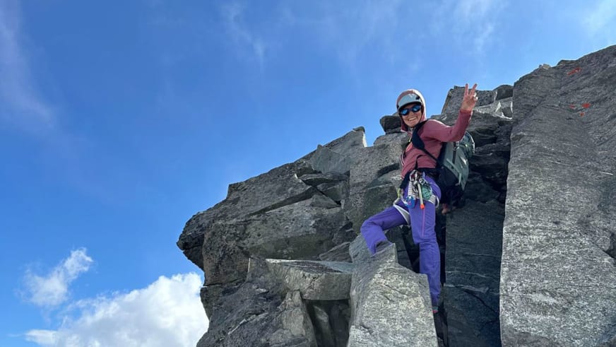 Mit weißem Helm und Klettergurt steht Bergführerin Elisabeth Seibt auf strukturiertem Fels und zeigt ein Victory-Zeichen gegen blauen Himmel.