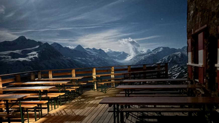 Leere Holzterrasse der Breslauer Hütte mit grünen Bänken vor schneebedeckter Bergkette der Ötztaler Alpen.
